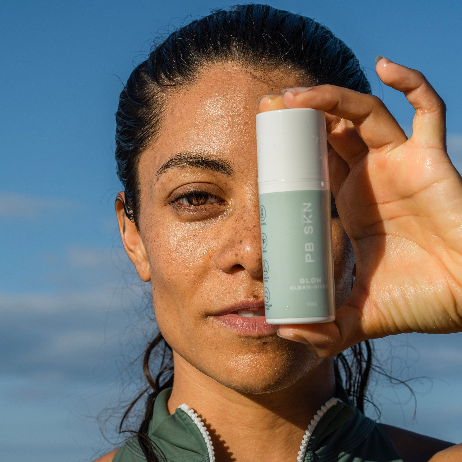 Woman holding PB SKN Glow brightening post-workout skincare serum at beach after exercise